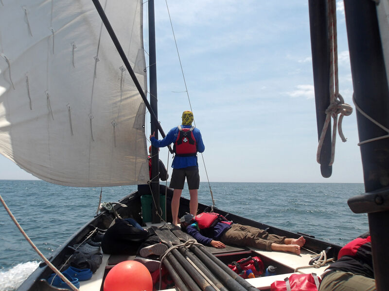 A sunny day on the water shows a sailboat cutting through the waves. A person in a blue shirt and yellow hat stands near the mast, looking out at the horizon. Another person relaxes on the deck, enjoying the ride. The boat's sail is full, catching the wind as it glides across the water.